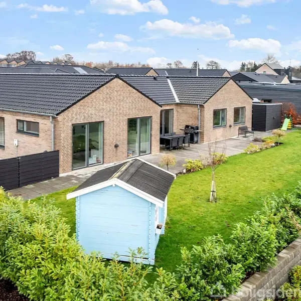 Et moderne murstenshus med store vinduer står ved en grøn have, hvor der er et lyseblåt skur. Haven er omgivet af en lav hæk, og der er en terrasse med havemøbler.