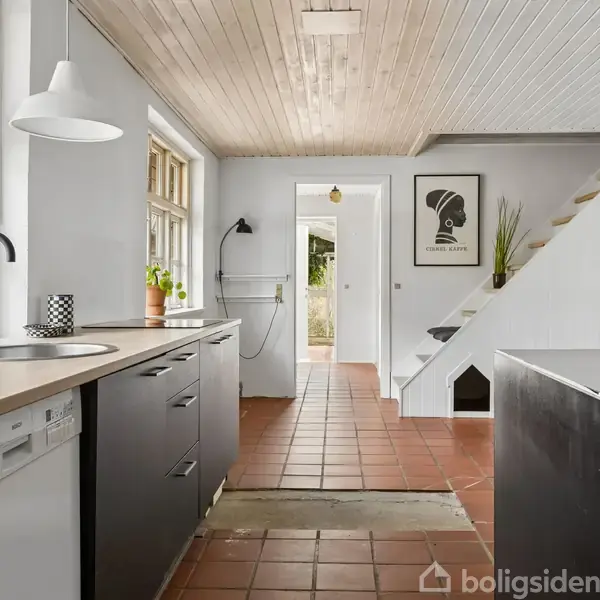 Køkken med sort-hvide skabe står i fokus. Vask og ovn er synlige, placeret langs bordpladen. I baggrunden ses trappe, plakat på væggen og planter under et træloft.