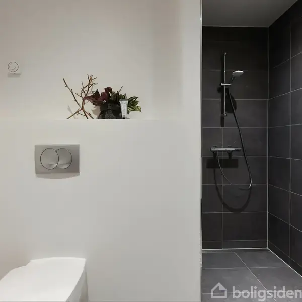 Toilet placeret i et lyst badeværelse, med en dekorativ plante ovenpå en separatorvægs hylde. Foran, til højre, ses en bruseniche med mørke fliser og moderne brusehoved.