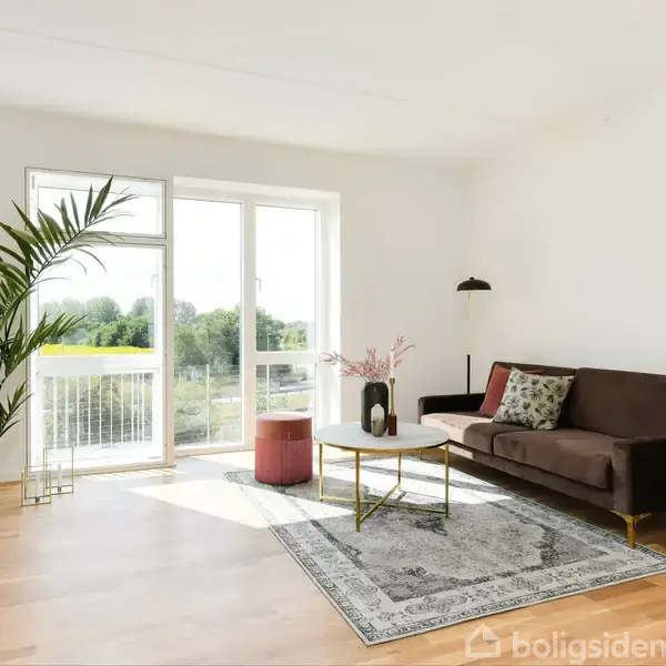 En stue med en brun sofa og et tæppe ligger ved et sofabord. En stor stueplante ved vinduet skaber en lys atmosfære. Ingen tekst er til stede.