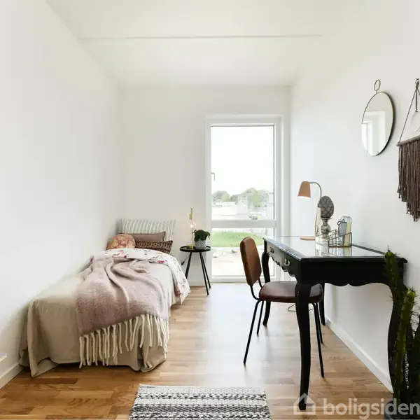 En enkeltseng dækket med tæpper står til venstre. Til højre er et skrivebord med en lampe og stol, nær et vindue, omgivet af minimalistisk indretning.