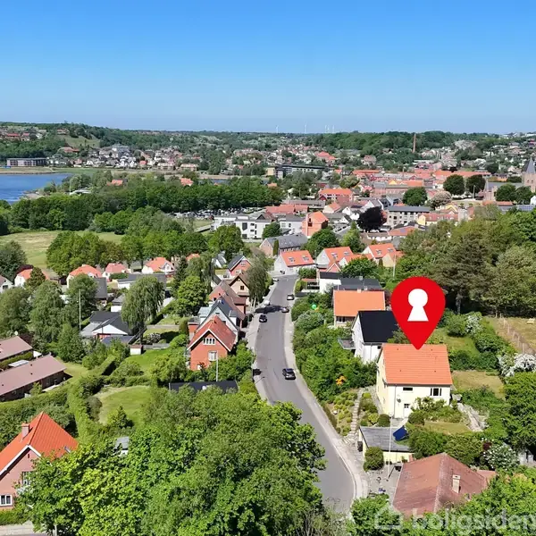 Hus markeret med en rød pil på taget i en bydel med grønne områder og spredte huse. Søen i baggrunden og kuperet terræn dominerer landskabet.