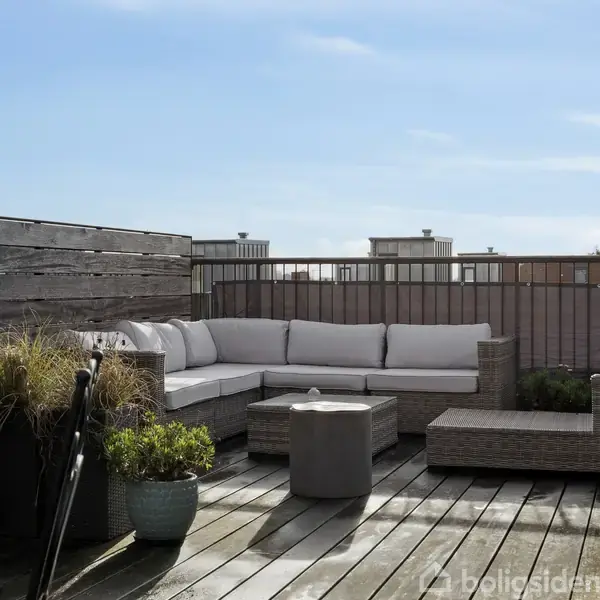 En hvid, hjørneorienteret udendørs sofa står på en træterrasse omgivet af planter i krukker og en lav trægærde, med en byudsigt i baggrunden.