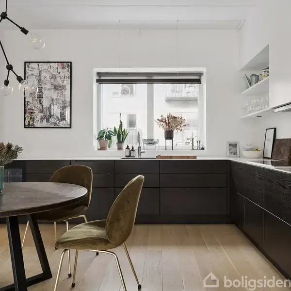 Et rundt træbord med fire stole står i et moderne køkken. Sorte skabe omkranser et vindue med planter og flasker på bordpladen. Lyse trægulve udgør rummets gulv.