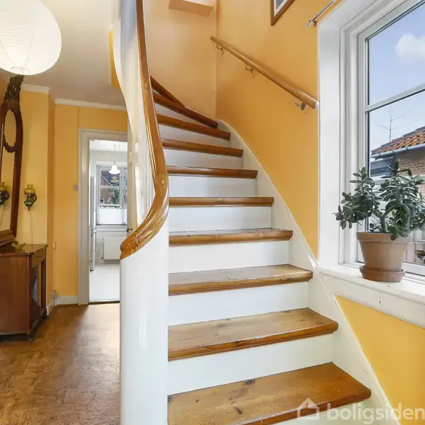 Trappe i træ fører opad ved siden af et lille vindue med en plante. Væggene er lysegule. Etageadskillelsen er synlig gennem en åbning.
