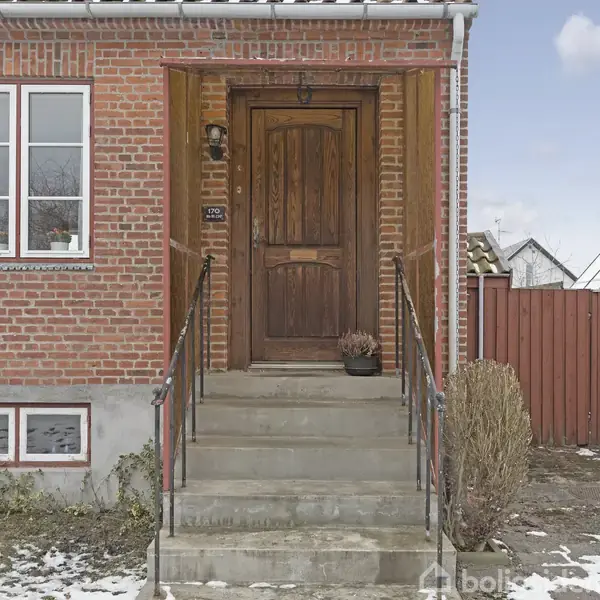 Rød murstensfacade med en trædør i midten, flankeret af to vinduer. Trappen leder til døren, omgivet af et gelænder. Der er snepletter på jorden foran huset. Husnummer: 170.