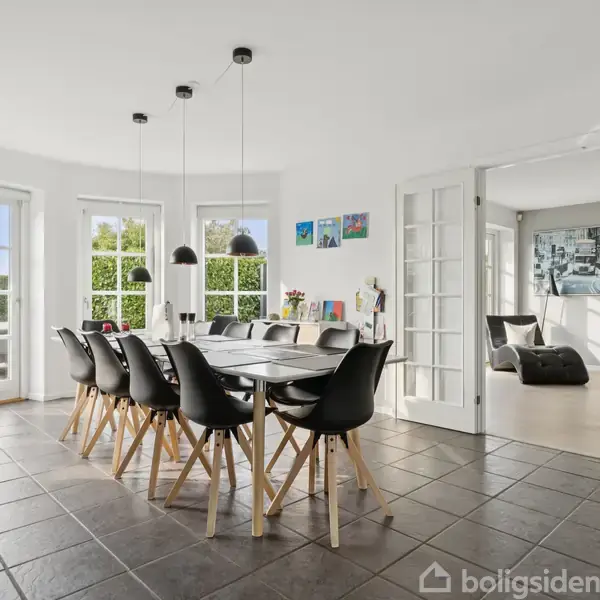 Spisebord med sorte stole omkring står under tre hængelamper. Store vinduer i lys spisestue med flisegulv og dør til tilstødende stue. Lyse, moderne omgivelser.