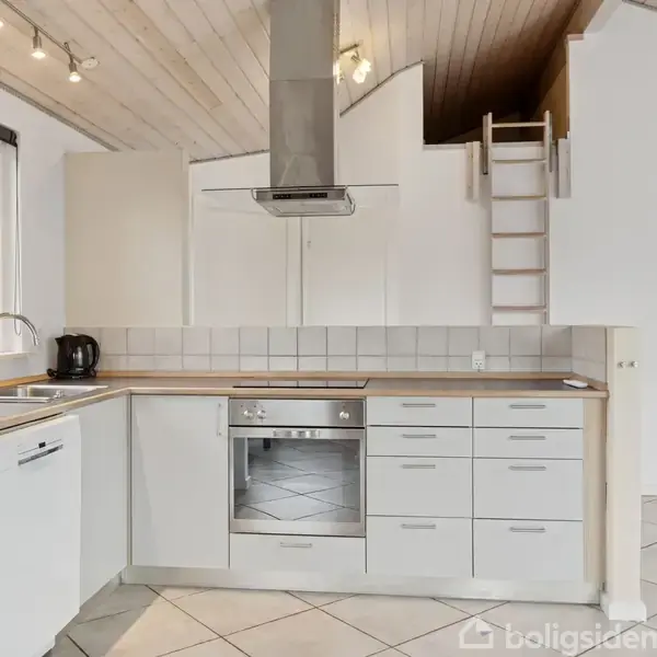 Køkken i lyse farver med hvidt køkkenskab, indbygget ovn og opvaskemaskine. Emhætte i rustfrit stål. Kedel ved vasken. Loft med træbeklædning og stige op til hemse. Flisegulv.