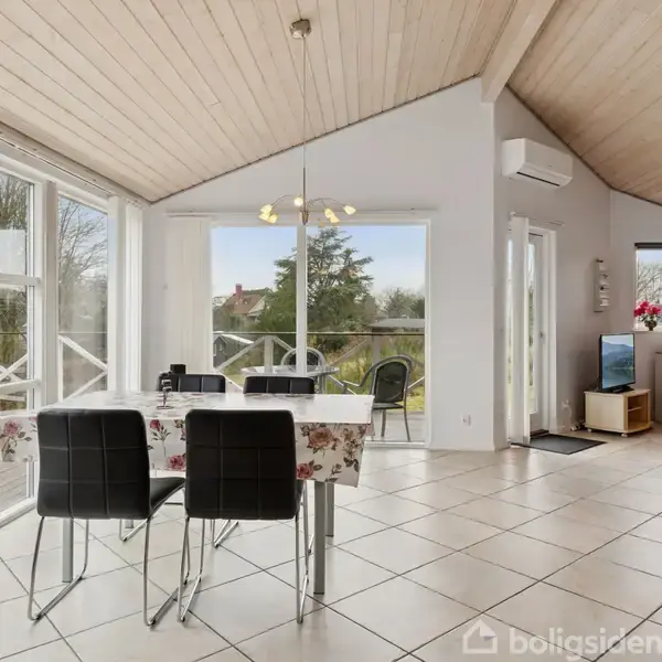 Spisebord med fire stole står i en lys stue med store vinduer og udsigt til terrasse. Loftet er træbeklædt, og der er en lampe over bordet. En computer står på et skrivebord i baggrunden.