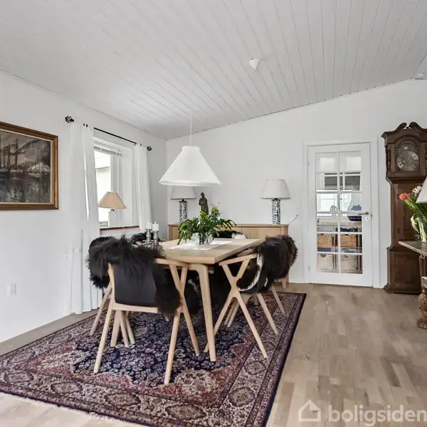 Et spisebord med seks stole står på et tæppe i en stue. Rummet er lyst med hvide vægge og træmøbler, herunder en kommode og et ur. Maleri og planter dekorerer rummet.