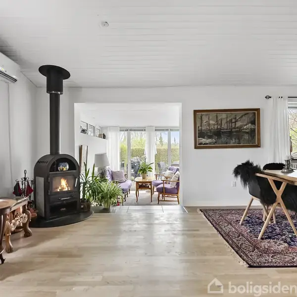 En brændeovn står i stuen og afgiver varme. Til venstre et rundt bord med blomster og stearinlys. Til højre et spisebord på et tæppe. Baggrund: vinduer med udsigt.