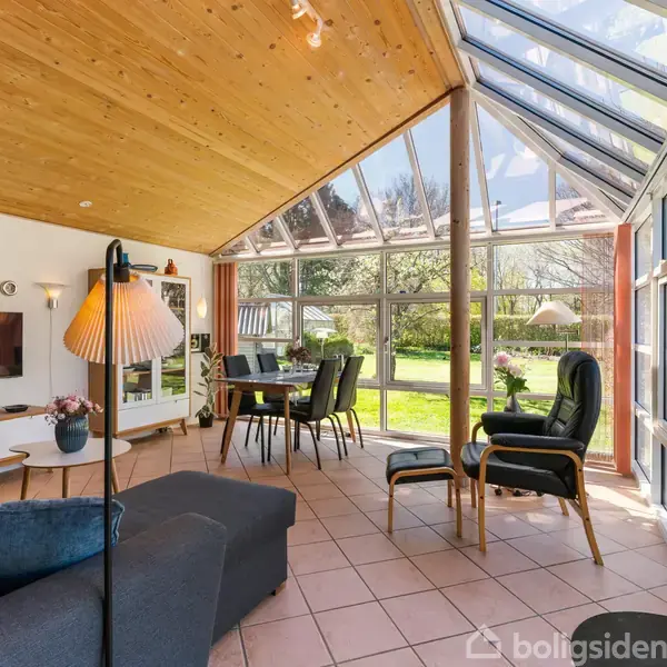 En stue med loft til kip og store glasvægge, indeholder en sofa, fjernsyn på væggen, spisebord med stole, og to lænestole; udsigt til en grøn have.