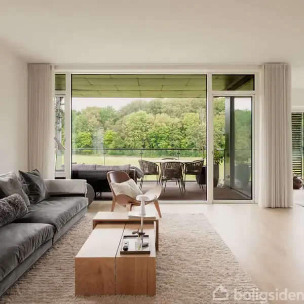 Stue med mørkegrå sofa og træbord står foran en stor, glasdækket terrasseudgang. Udenfor er der et spisebord og en udsigt over grønne træer.