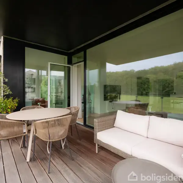 En udendørs terrasse med en sofa og rundt bord, omgivet af glasvægge og grønne planter. Indenfor ses træer og en afslappet stue.