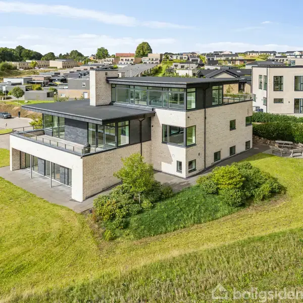 Et moderne hus med store vinduespartier og flade tage står på en grøn bakke. I baggrunden ses en række huse, der danner en boligkvarter.
