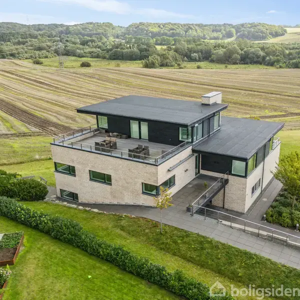 Et moderne hus med fladt tag og store vinduer ligger ved siden af en solrig terrasse, omgivet af velholdt græsplæne og bølgende marker i en bakket landskab.