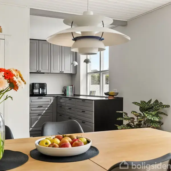 En spisebord med en skål frugt og en vase med blomster står centralt. En moderne lampe hænger ovenover. I baggrunden ses et køkken med grå skabe og lysindfald fra vinduer.
