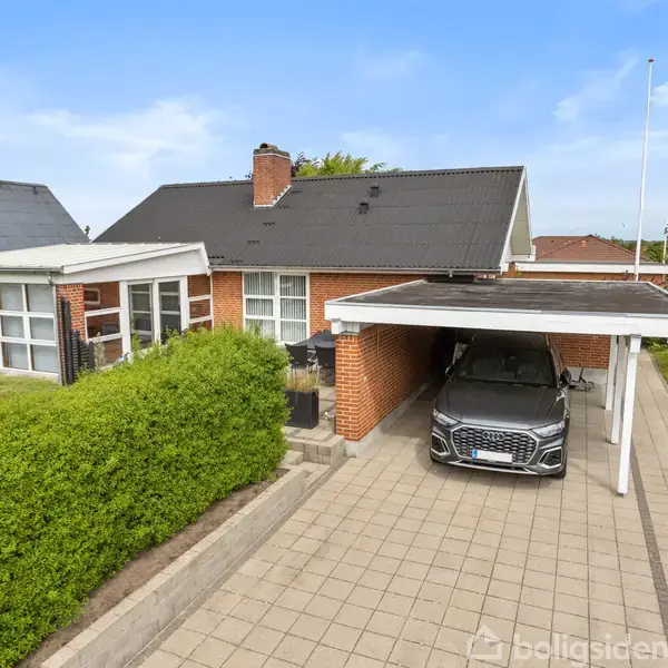 Et rødstenshus med sort tag har en tilknyttet carport, hvor en grå bil er parkeret. Hækken omkranser indkørslen, og en flisebelagt sti fører til hovedindgangen.