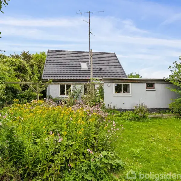 Hus med gråt tag står i en frodig have med græsplæne og blomsterbede. En antenne er monteret på taget. Omgivet af grønne planter og træer.