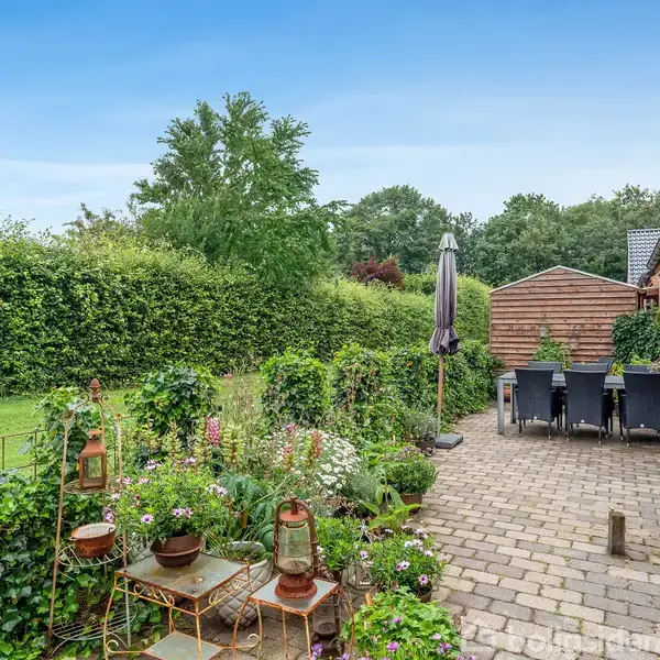 Terrasse med fliser fyldt med blomsterkrukker og lanterner. Et bord med stole og en parasol står langs husmuren. Omringet af hække og træer i en grøn have.