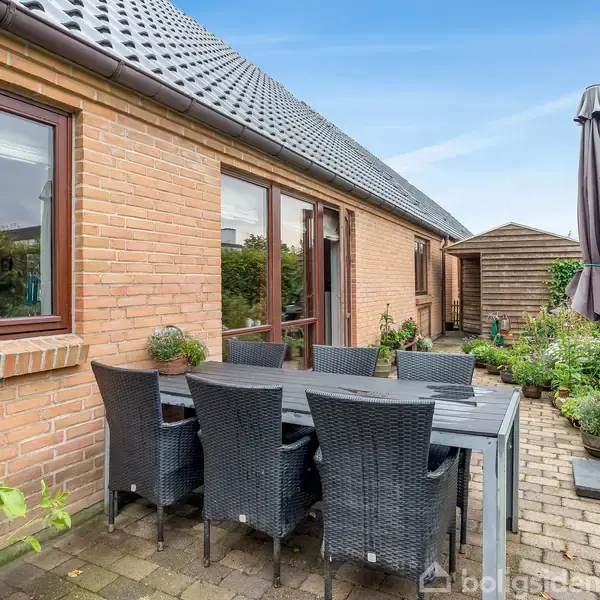 En sort spisebord med seks fletstole står på en brolagt terrasse foran et murstenshus. Planter og en parasol omkranser området, der ligger op ad en have.