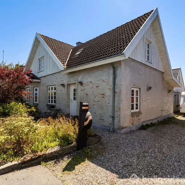 Et gråhvidt murstenshus med skråt tag står ved en grusbelagt indkørsel. En person går foran huset, omgivet af en have med buske. En campingvogn er parkeret ved siden af.
