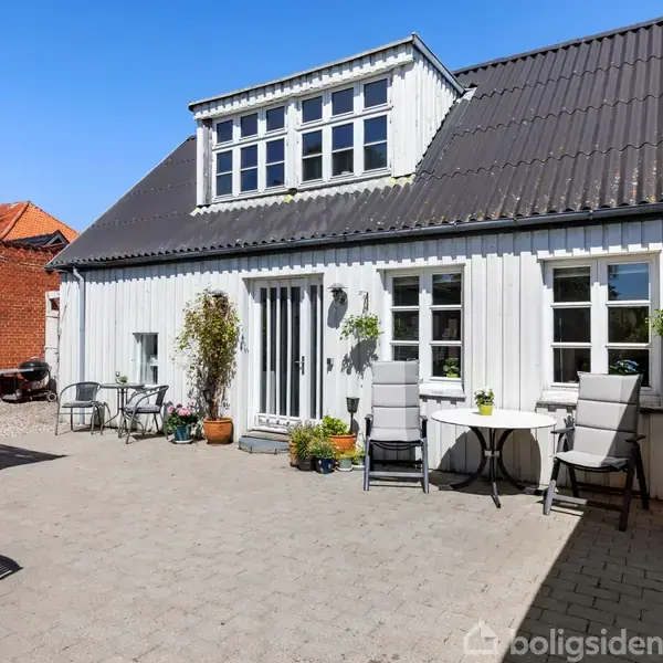 Hvidt hus med mørkt tag står i en gårdhave fyldt med havemøbler og blomsterkrukker. Blå himmel i baggrunden.