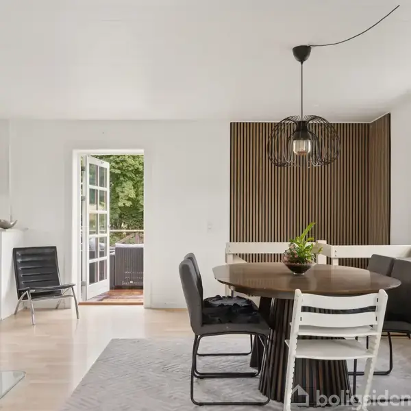 En spiseplads med et rundt bord og forskellige stole står i en lys stue. Døren i baggrunden åbner ud til en terrasse, og et stort vindue giver udsigt til haven. En moderne pendellampe hænger over bordet.