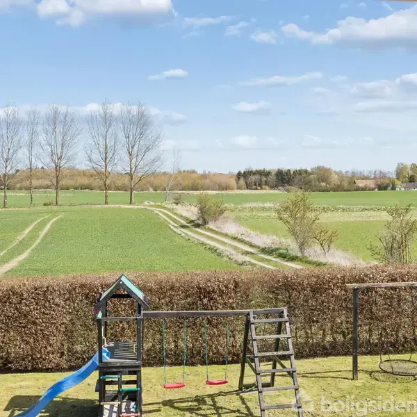 Legeplads med gynger og rutsjebane står stille i en have, omkranset af hække. I baggrunden ses åbne marker og en række træer under en blå himmel med få skyer.