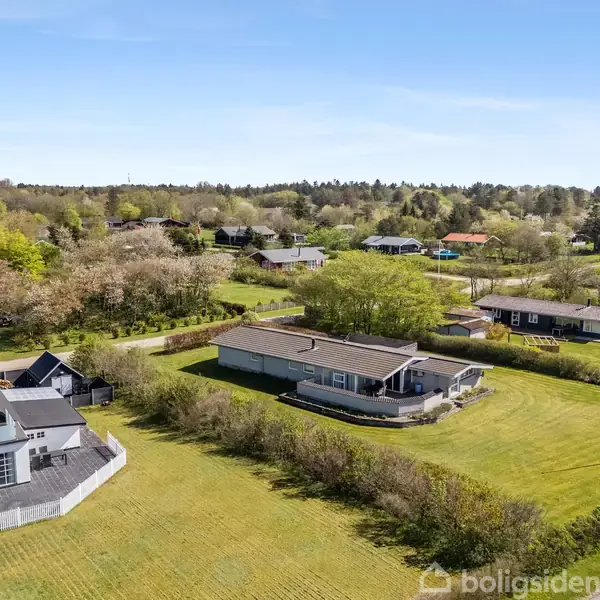 Et moderne hus med fladt tag er placeret på en stor græsplæne omgivet af buske. I baggrunden ses flere huse spredt i et grønt, skovfyldt landskab.