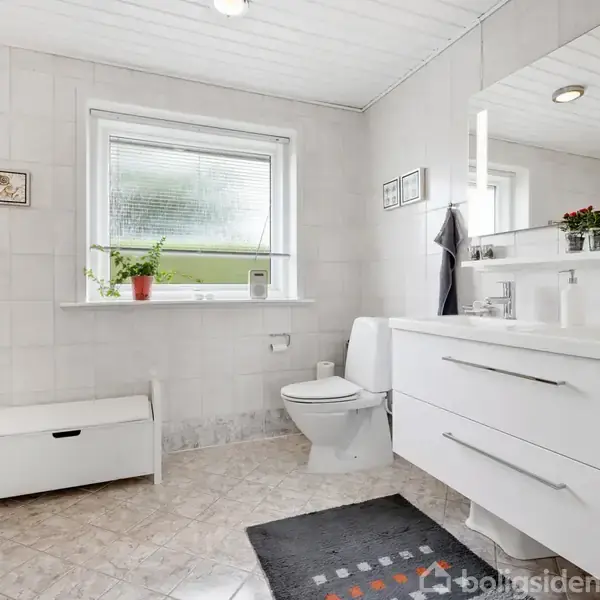 Badeværelse med hvidt skab og vask, toilet, og vindue med planter. Håndklæder hænger ved spejlet. Lyse fliser på vægge og gulv. Lille bænk står ved siden af toilettet.