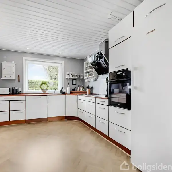 Moderne køkken med hvide skabe og trægulve, to indbyggede ovne. Et stort vindue slipper naturligt lys ind. Grønne planter dekorerer rummet.