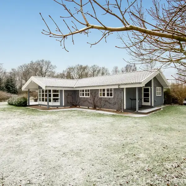 Et gråt træhus står stille på en snedækket plæne, omringet af træer og buske, under en klar, blå himmel.