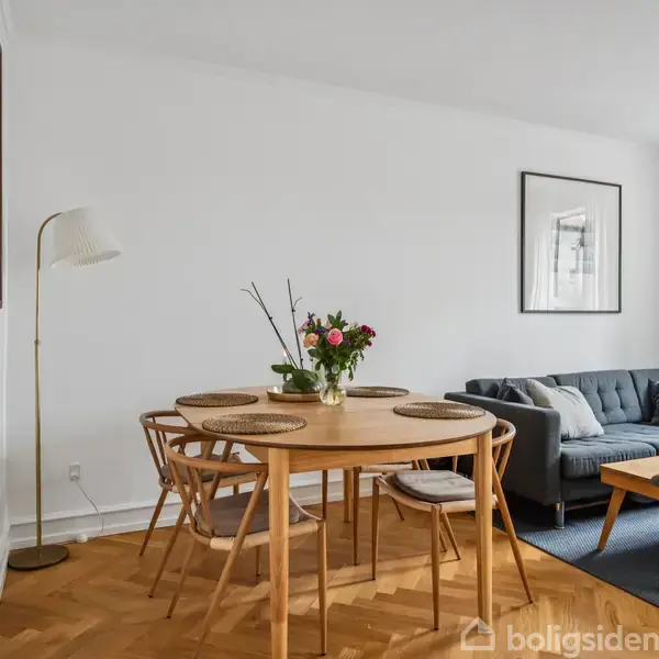 Rundt spisebord med fire stole, pyntegenstande og blomster. I baggrunden en grå sofa og et vægmaleri. Rummet har trægulv og hvidmalede vægge. Ingen tekst er synlig.