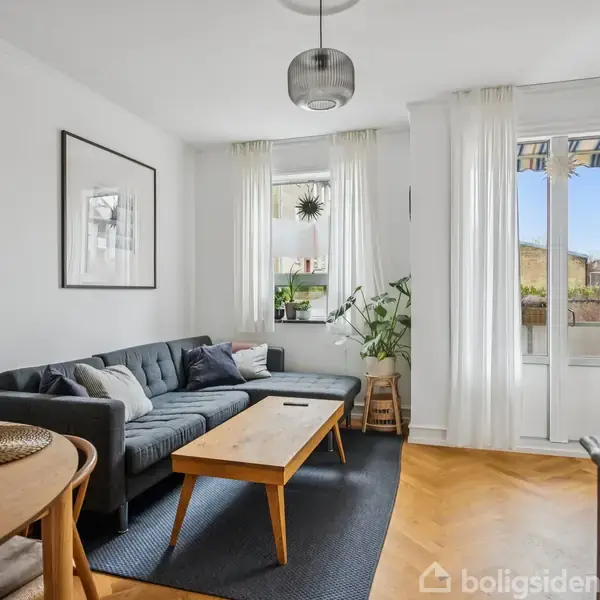 Sofaen står mod væggen i en lys stue med trægulv, hvor et sofabord står foran. I baggrunden er et vindue og glasdør med udsigt til en terrasse.