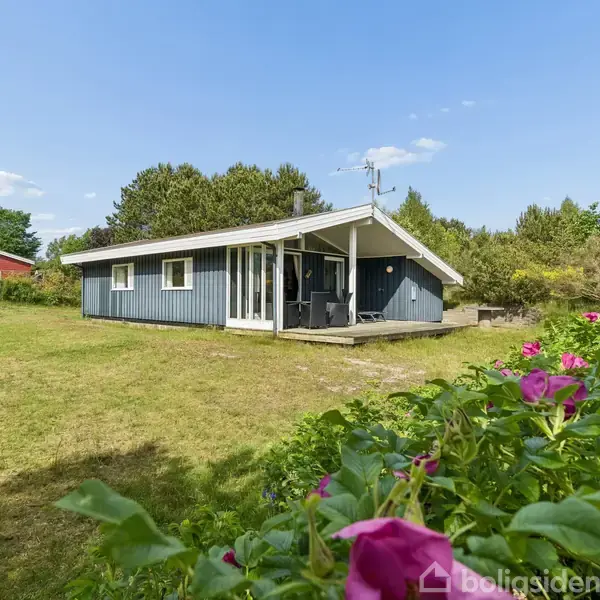 Blåt træhus står stille i en grøn have med rosa blomster i forgrunden. Det er omgivet af buske og træer under en blå himmel med få skyer.