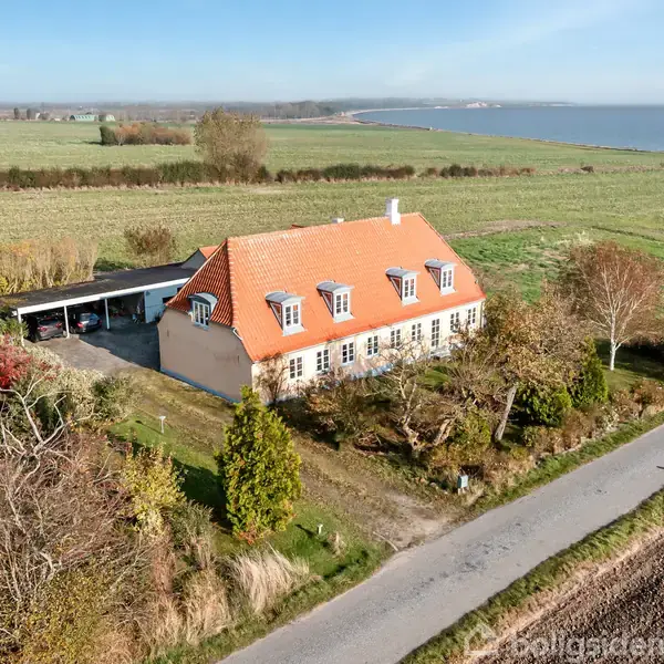 Et landhus med rødt tegltag og hvide vinduer står ved en vej. Det er omgivet af træer og buske, med marker og hav i baggrunden.