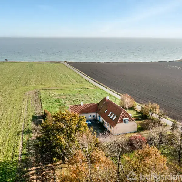 Hus med rødt tag ligger omgivet af marker, tæt på kysten med havudsigt. Omkring huset er træer og en vej, der løber langs en brun mark.