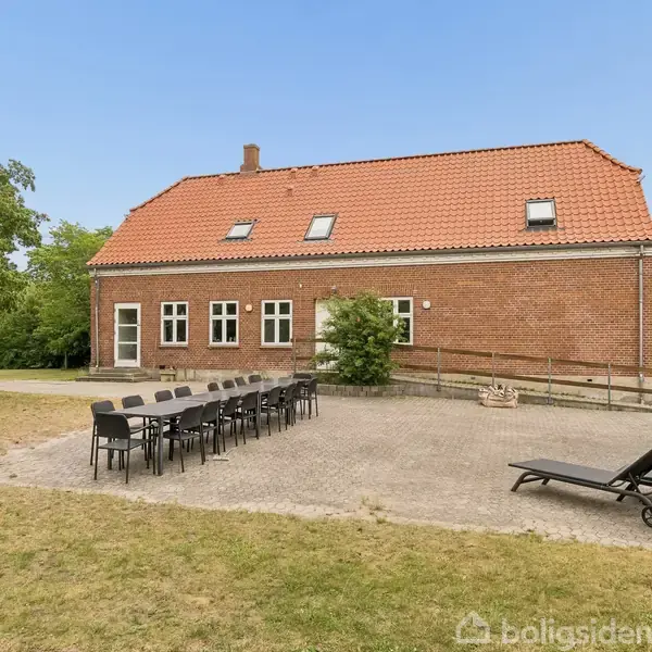 Rødstenshus med rødt tegltag og vinduer, omgivet af græs og træer. Langt bord med sorte stole og solsenge på en flisebelagt terrasse foran huset.