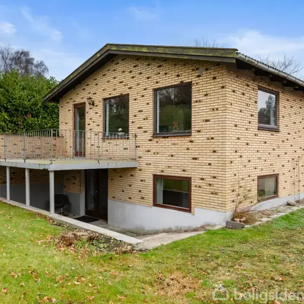 Et gulstenshus står udenfor med en hævet terrasse. Græsplæne og buske omgiver huset under en klar himmel.