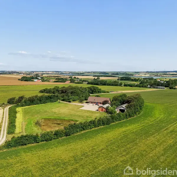 Gård beliggende midt i store grønne marker med skovklædte områder omkring. En grusvej fører ind til gården. Landskabet er fladt med udsigt over landbrugsjord under en blå himmel.