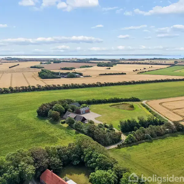 En gård med bygninger omgivet af skov arealer. Gården befinder sig i et åbent landskab med opdyrkede marker, der strækker sig mod horisonten under en blå himmel med lette skyer.