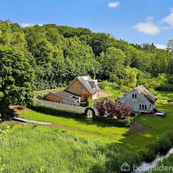 To huse står i et grønt, skovfuldt landskab. Det ene hus er gult med en mørk tag, og det andet er gråt. En lille å løber gennem grunden.