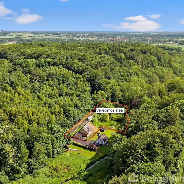 En bygning er omringet af tæt skov. Skiltet "Vejledende areal" markerer grunden. Omgivet af frodigt landskab med marker og træer i det fjerne under en let overskyet himmel.