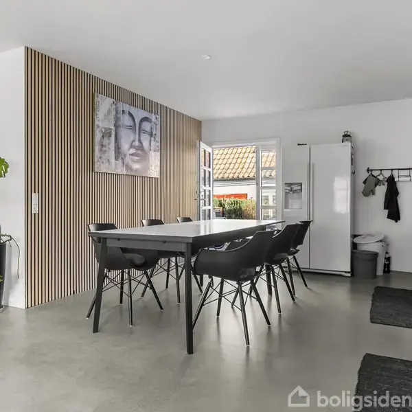 Et moderne køkken-alrum med et langt spisbord og seks sorte stole. En plante står til venstre, og kunstværker hænger på væggen. Et hvidt køleskab står ved en bagdør.