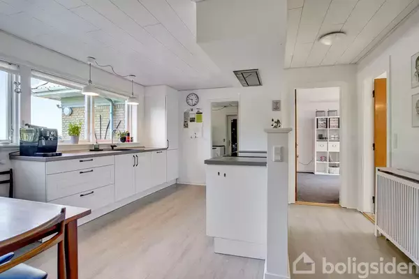 Et lyst køkken med hvide skabe og sort bordplade, hvor en kaffemaskine står ved vinduerne. Spisebord og stol ses til venstre. En dør forbinder til en anden stue.