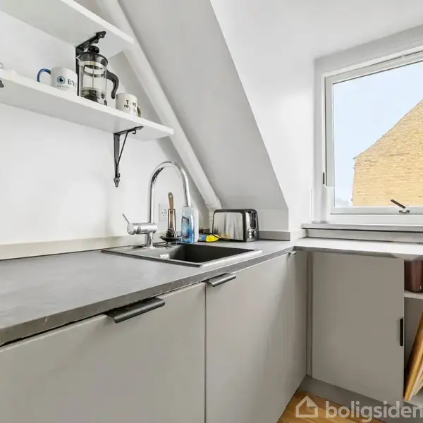 Et lille, lyst køkken med hvide skabe, en sort håndvask og bordplade. Hylder over vasken indeholder køkkenudstyr. Et vindue til højre giver udsigt til en murstensbygning.