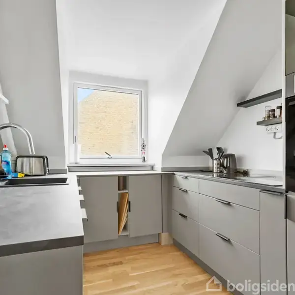 Et moderne køkken med grå skabe og skuffer står ubenyttet i et lyst rum med trægulv. En vask med vandhane er placeret under et stort vindue, og en ovn ses til højre.