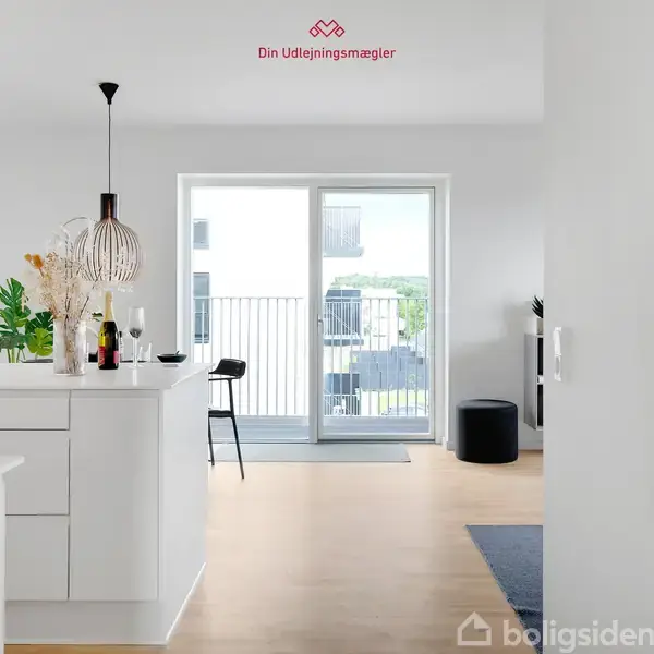 Et moderne køkken med hvide skabe og en central ø, dekoreret med planter og flasker. I baggrunden ses en stor altandør, der fører ud til en balkon. Tekst: "Din Udlejningsmægler".