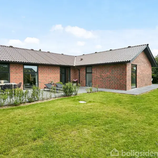 Huset er en moderne, rød murstensvilla med sorttag, omgivet af en frodig have med anlagt plæne. Terrasse med borde og stole ses foran huset, som står i solskin.